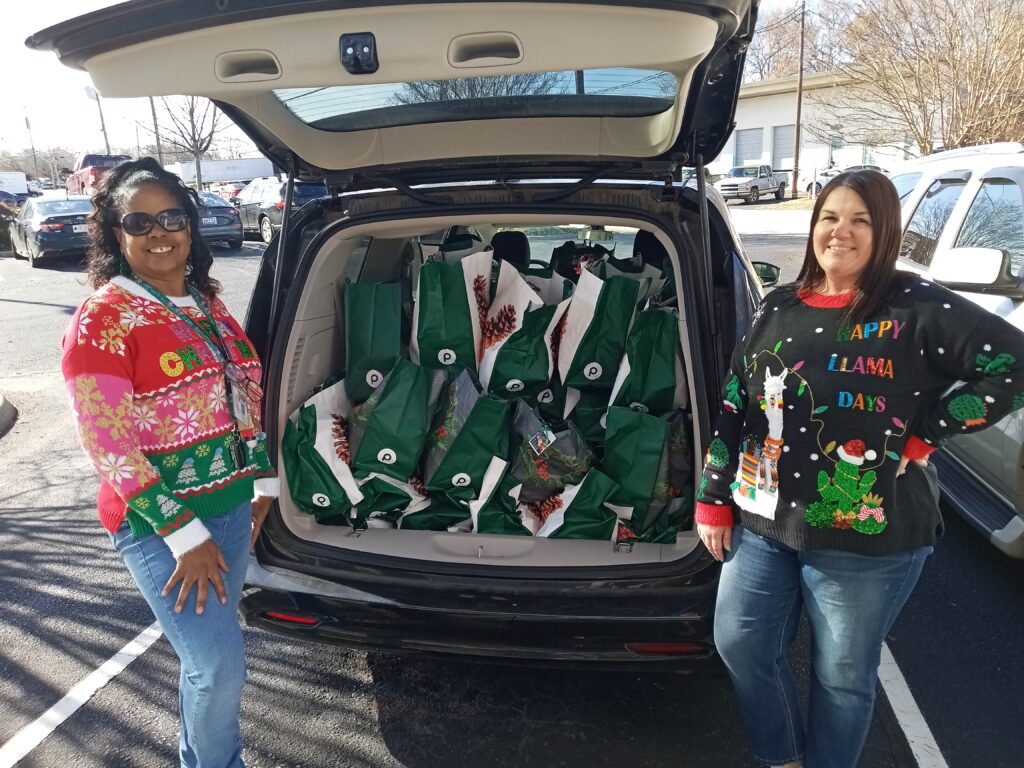 A black woman in a christmas sweater and sunglasses, and a white woman in a black Christmas sweater smile in front of an open minivan loaded up with green and white Publix bags printed with pinecones.