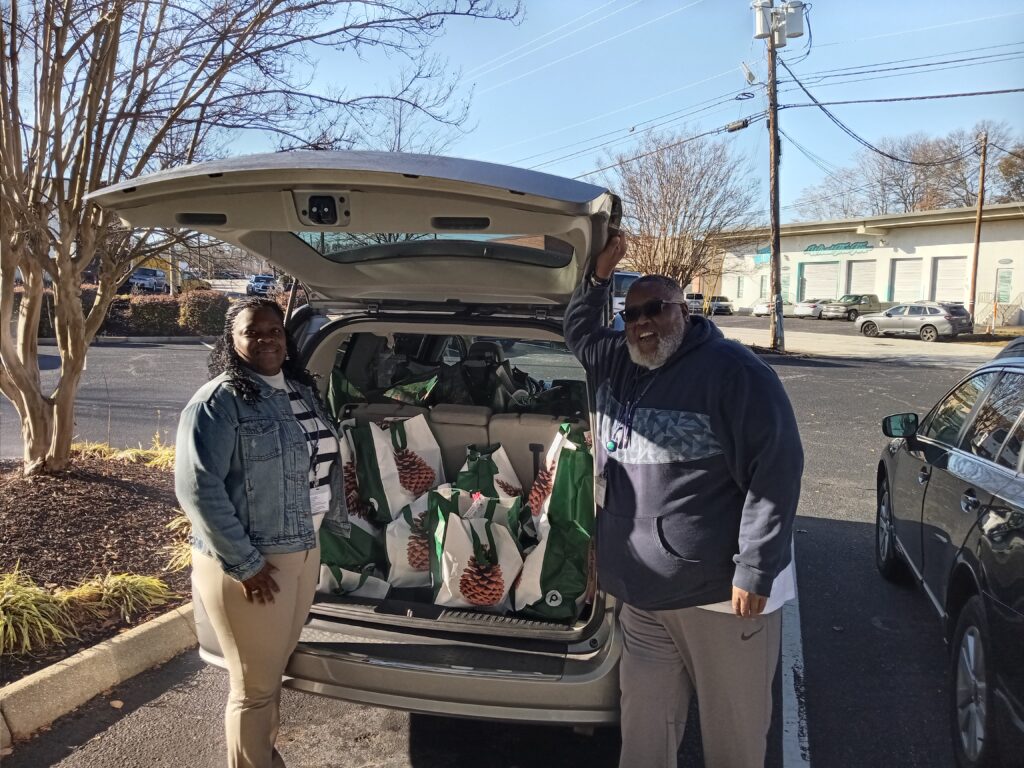 A black woman in a striped shirt and a jean jacket and a black man wearing a navy hoodie and sunglasses stand in front of an open minivan that is full of green and white publix bags printed with a pine cone.