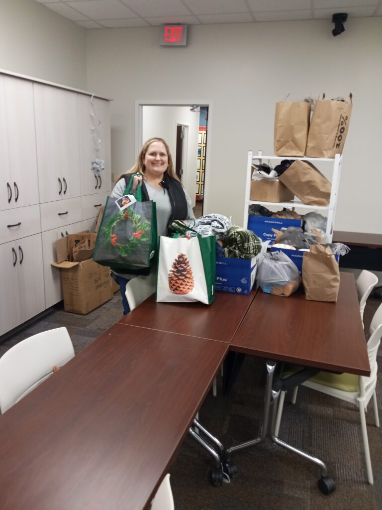 A smiling white woman with long blond hair stands at the end of a table covered in festive bags and boxes loaded with blankets.