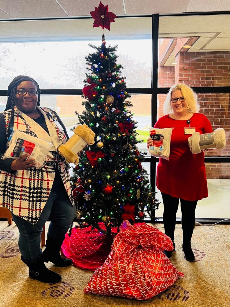 A black woman in a plaid holiday wrap and a white woman in a red dress stand on opposite sides of a decorated christmas tree. They hold personal care items.