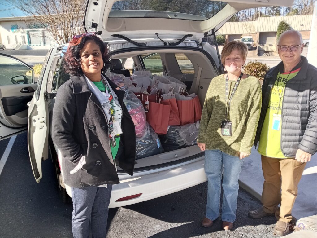 A black woman in a grey jacket and a chrismas shirt stands to the left of an open mini van. A white woman in a green sweater and a white man in a Grinch shirt and grey puffy jacket stand on the other side. The back of the van is full of holiday bags.