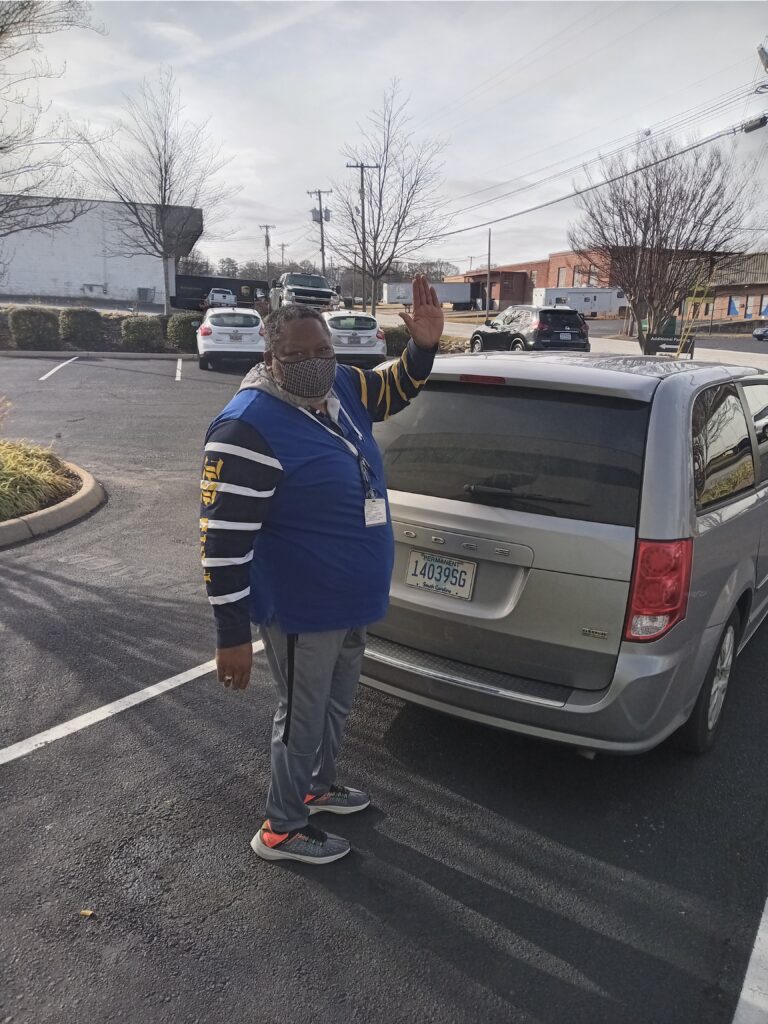 A black man in a blue vest and face mask stands beside a minivan waving at the photo taker.