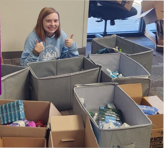 A young girl with red hair, wearing a blue hoodie smiles and gives two thumbs up, surrounded by grey boxes full of toiletry items.