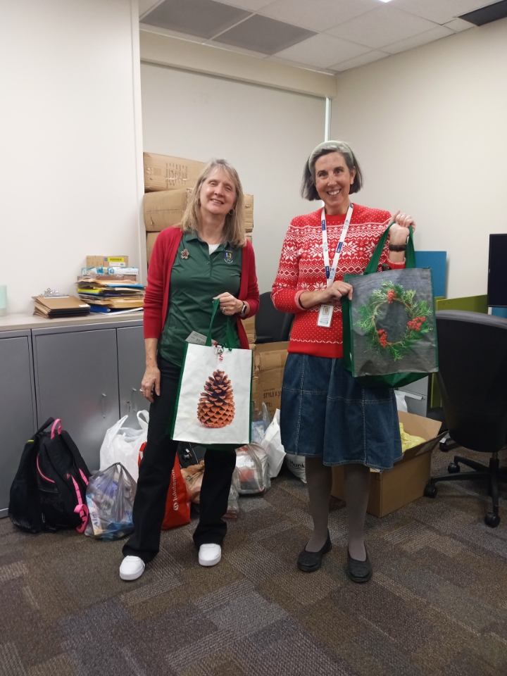 Two white women in Christmas colors stand in an office, each holding a christmas themed shopping bag.