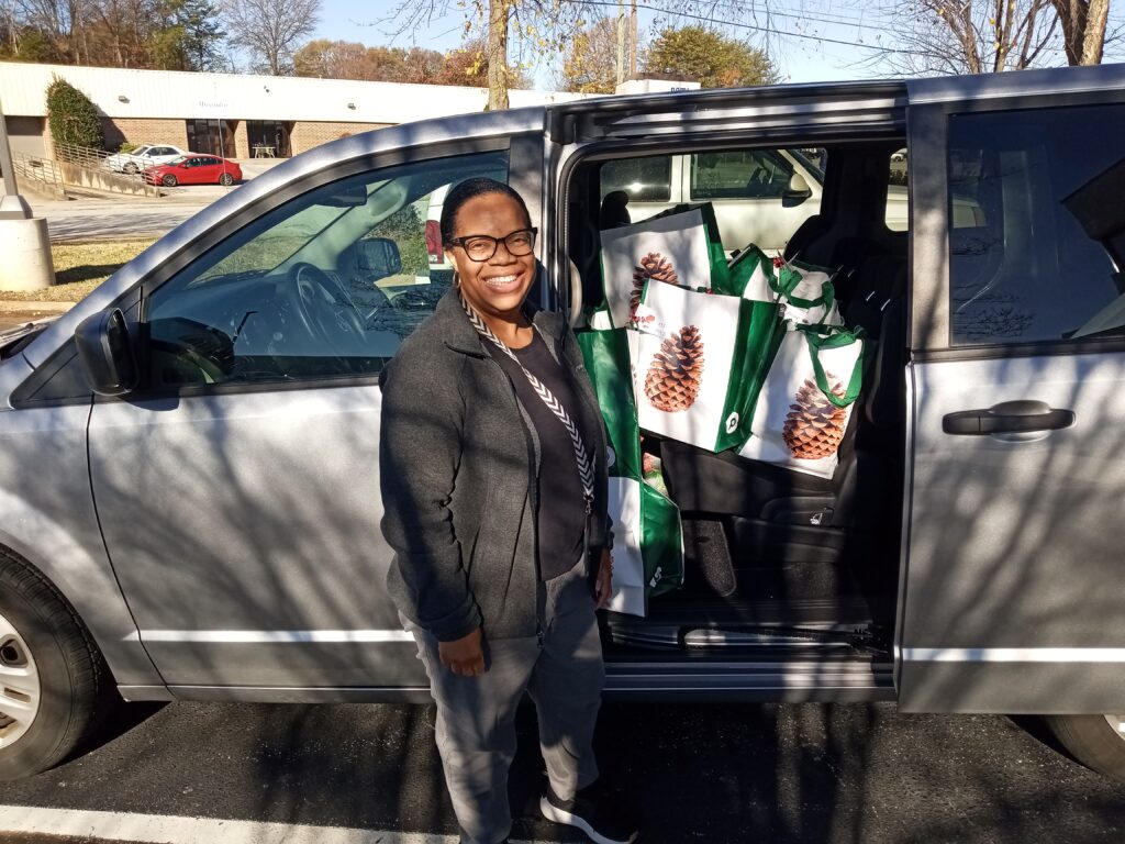 A black woman wearing a dark grey coat and glasses stands beside an open minivan. The back seat is full of green and white publix bags with Pinecones printed on them.