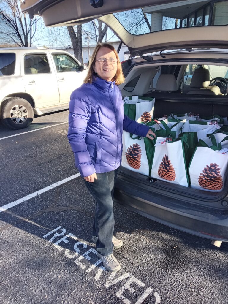 A blond woman in glasses wearing a purple jacket stands infront of the open hatch of a minivan. The back is full of white bags with Pinecones printed on them.