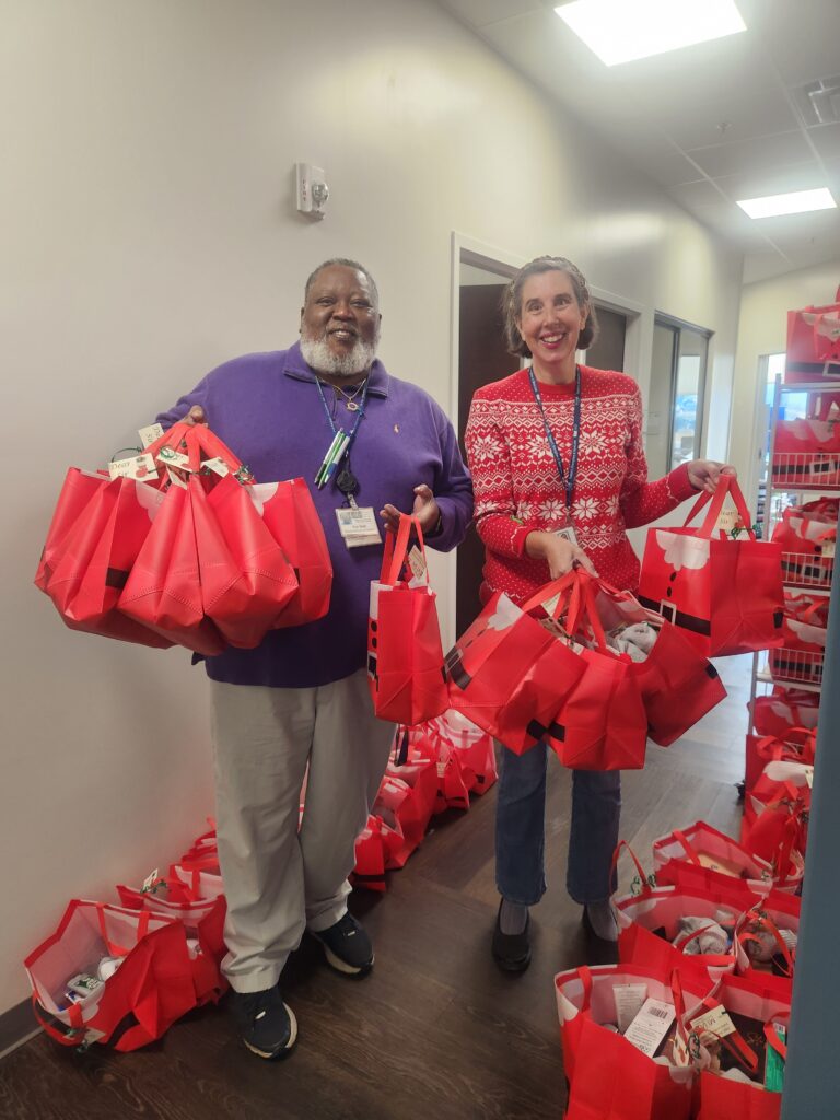 A smiling man and woman stand in a hall way filled with holiday bags, They both have their arms full of the holiday bags.